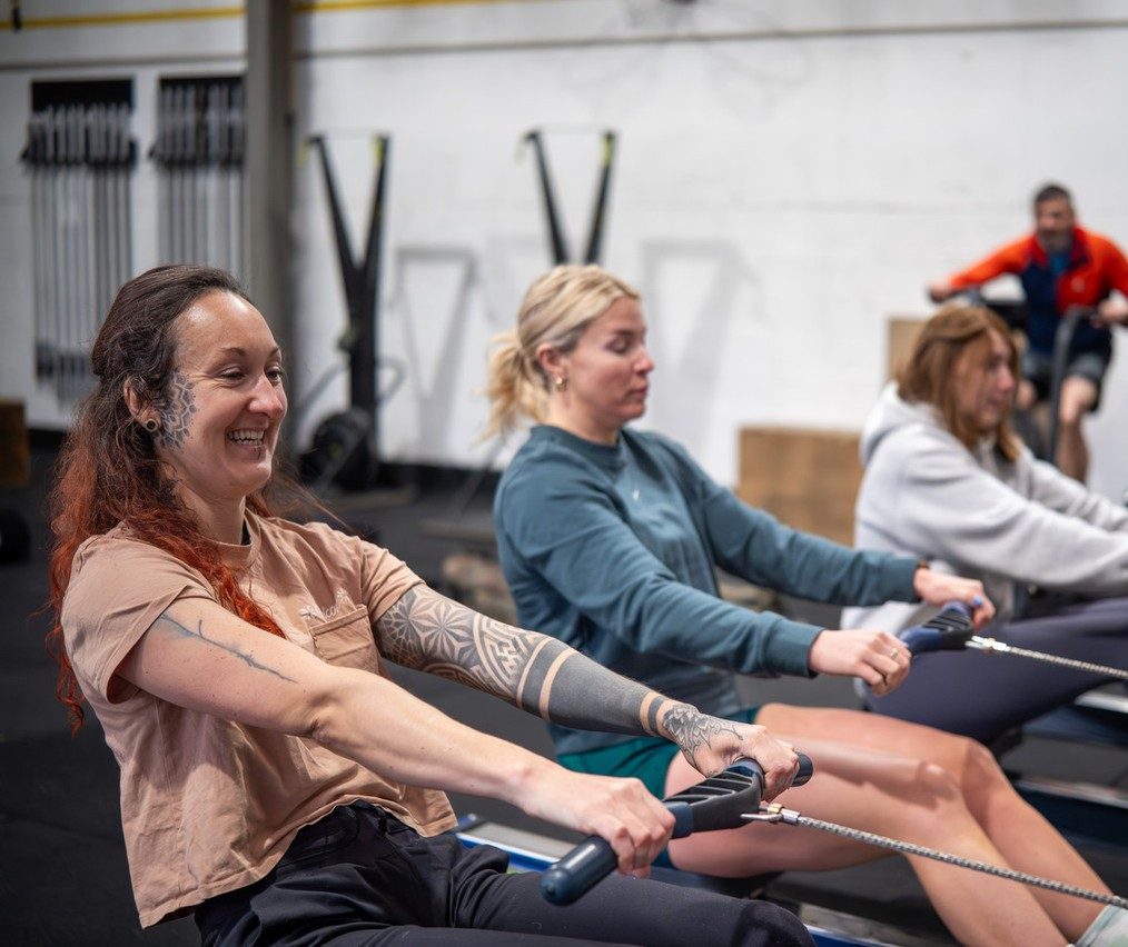 Three CrossFit Bodmin members smiling on the rowers during a coached class